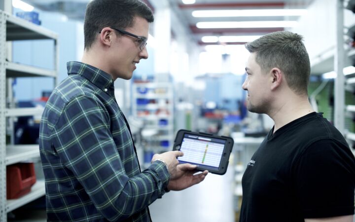 Mitarbeiter aus Büro und Fertigung unterhalten sich vor Tablet mit Inhalten der Fertigung